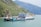 A large ferry boat is carrying several trucks across a wide river. The boat has a multi-story structure and is painted in shades of blue and white. There are green, hilly terrains on the left side and mountainous landscapes in the background with a calm sky.