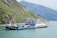 A large ferry boat is carrying several trucks across a wide river. The boat has a multi-story structure and is painted in shades of blue and white. There are green, hilly terrains on the left side and mountainous landscapes in the background with a calm sky.