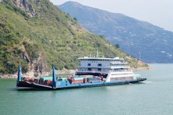 A large ferry boat is carrying several trucks across a wide river. The boat has a multi-story structure and is painted in shades of blue and white. There are green, hilly terrains on the left side and mountainous landscapes in the background with a calm sky.