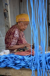 An elderly person wearing a yellow headscarf and a patterned red blouse is sitting and working with their hands. In front of them is a stack of coiled blue rope. The background features a wooden structure and a hanging object, possibly a tool or container.