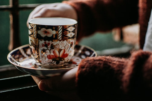 A person's hands hold a beautifully decorated porcelain teacup and saucer. The teacup features intricate floral and geometric patterns with vibrant colors, primarily red, black, and gold, against a softly blurred background.