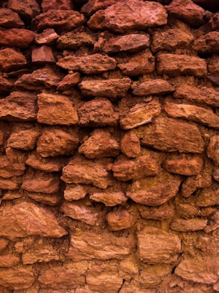A close-up view of a textured stone wall composed of various sizes and shapes of reddish-brown rocks. The stones are stacked in an uneven pattern, showing a natural and rustic appearance.