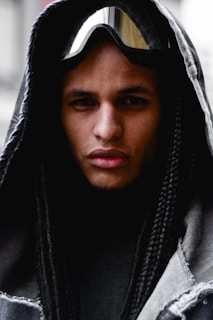 A person is wearing a hooded garment with long, braided hair and reflective goggles resting on their forehead. The hood is made of a dark material that frames their face closely, and the goggles add a futuristic or adventurous element to the look.