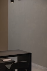 A minimalist and modern interior setting with a dark table and chair positioned against a plain, muted wall. A small white book or notepad rests on the table. Overhead, a black cylindrical light fixture hangs from the ceiling, contributing to the subtle ambiance.