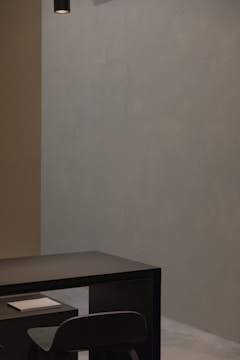 A minimalist and modern interior setting with a dark table and chair positioned against a plain, muted wall. A small white book or notepad rests on the table. Overhead, a black cylindrical light fixture hangs from the ceiling, contributing to the subtle ambiance.