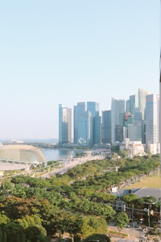 A vibrant cityscape with modern skyscrapers in the background, surrounded by lush greenery and a prominent waterfront area. The architecture includes a unique, rounded building and a clear blue sky enhances the scene.