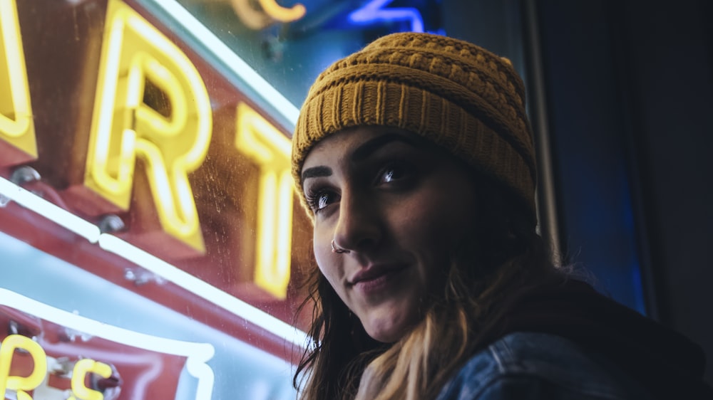 woman wearing yellow beanie while standing near neon light sign