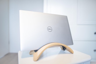 A silver Dell laptop placed on a wooden stand, situated on a white table against a light-colored wall. The scene is minimalistic and clean, with the focus on the sleek design of the laptop and its stand.