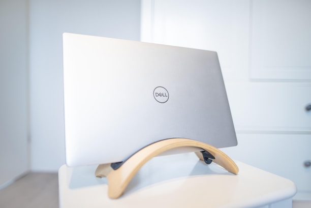 A silver Dell laptop placed on a wooden stand, situated on a white table against a light-colored wall. The scene is minimalistic and clean, with the focus on the sleek design of the laptop and its stand.