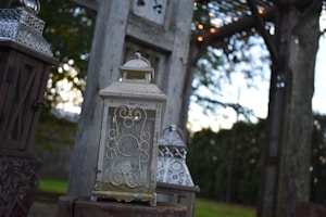 A decorative outdoor setting features ornate lanterns placed on wooden stumps. The backdrop includes a rustic wooden structure and greenery, with soft lighting creating a cozy atmosphere.