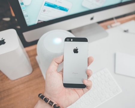 A hand holding a silver iPhone with the Apple logo visible, in a workspace setting. Surrounding items include a large monitor, a wireless keyboard, and a smart speaker. The desk is made of wood and has a minimalist aesthetic.