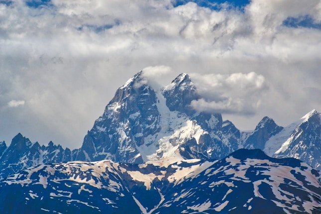 A majestic mountain range with snow-capped peaks under a cloudy sky. The central peak is prominent and partially shrouded by clouds, with rugged slopes contributing to the dramatic landscape. The foreground is covered with patches of snow, contrasting against darker rocky areas.