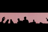 Silhouetted figures are positioned against a light pink rectangle backdrop, suggesting a social setting. There are multiple people visible, with subtle details indicated by the outlines. In the foreground, a hand gesture is noticeable, possibly suggesting a conversational or lively interaction.