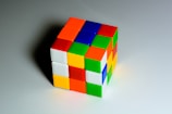 A partially solved Rubik's Cube is positioned on a flat, light-colored surface. Each side of the cube features a grid of smaller squares with different colors, including red, blue, green, yellow, orange, and white.