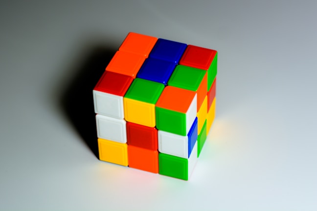 A partially solved Rubik's Cube is positioned on a flat, light-colored surface. Each side of the cube features a grid of smaller squares with different colors, including red, blue, green, yellow, orange, and white.
