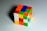 A partially solved Rubik's Cube is positioned on a flat, light-colored surface. Each side of the cube features a grid of smaller squares with different colors, including red, blue, green, yellow, orange, and white.