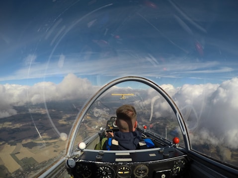 A view from inside the cockpit of a glider, showing the back of the pilot's head. The glider is flying among large, fluffy clouds with a clear blue sky above. Below, patches of farmland can be seen. An instrument panel is visible in the foreground, and another aircraft is flying ahead at a distance.