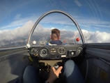The view from inside a cockpit, showing an individual piloting a glider or light aircraft. The dashboard contains various gauges and instruments, and the sky outside is bright with expansive clouds. The pilot is wearing a blue jacket and holds a control stick.