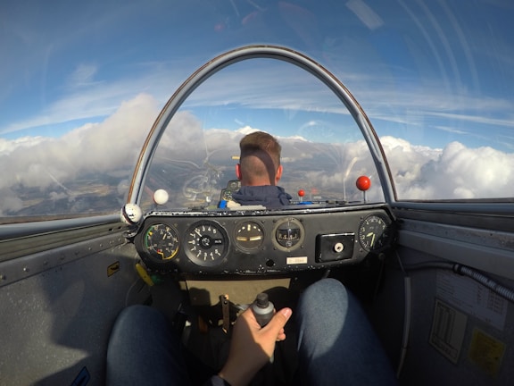 The view from inside a cockpit, showing an individual piloting a glider or light aircraft. The dashboard contains various gauges and instruments, and the sky outside is bright with expansive clouds. The pilot is wearing a blue jacket and holds a control stick.