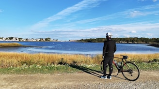 A person dressed in black cycling attire stands beside a bicycle on a gravel path. They face a serene body of water with a clear blue sky overhead. The landscape features tall grasses in the foreground and a distant view of trees and buildings on the horizon.