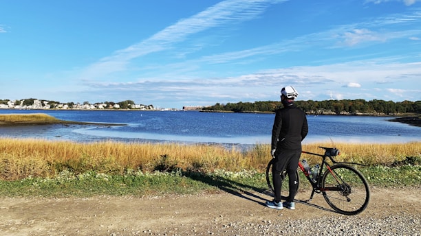 A person dressed in black cycling attire stands beside a bicycle on a gravel path. They face a serene body of water with a clear blue sky overhead. The landscape features tall grasses in the foreground and a distant view of trees and buildings on the horizon.