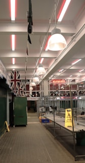 An indoor market with metal stalls and bright overhead lights. The walls are adorned with large Union Jack flags, and there are warning signs placed around indicating potential hazards. The market appears empty and quiet, with no people present.