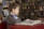 A young boy seated at a red table engrossed in a large, open book. Surrounding him are shelves filled with colorful books, suggesting a library or bookstore setting. His expression is focused and absorbed.