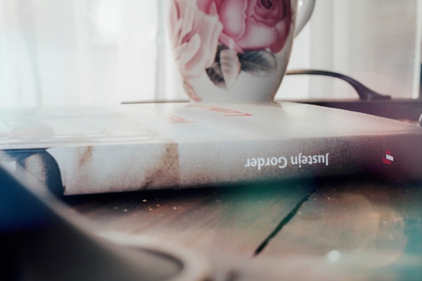 A hardcover book is lying on a wooden surface with a floral mug placed next to it. The cover of the book has a textured appearance, and the name 'Justeïn Cordero' is visible on the spine. The mug is decorated with pink roses and green leaves. The background is softly lit, giving a cozy and intimate feel.