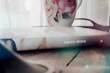 A hardcover book is lying on a wooden surface with a floral mug placed next to it. The cover of the book has a textured appearance, and the name 'Juste&iuml;n Cordero' is visible on the spine. The mug is decorated with pink roses and green leaves. The background is softly lit, giving a cozy and intimate feel.