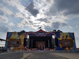 A large outdoor stage decorated with a carnival theme, featuring colorful graphics of balloons and a carousel. The stage is set under a cloudy sky with sunlight filtering through the clouds. Branding elements, such as a Pepsi logo, are prominently displayed.