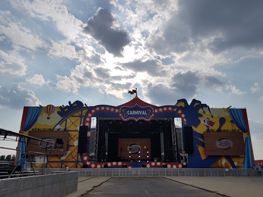 A large outdoor stage decorated with a carnival theme, featuring colorful graphics of balloons and a carousel. The stage is set under a cloudy sky with sunlight filtering through the clouds. Branding elements, such as a Pepsi logo, are prominently displayed.