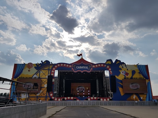 A large outdoor stage decorated with a carnival theme, featuring colorful graphics of balloons and a carousel. The stage is set under a cloudy sky with sunlight filtering through the clouds. Branding elements, such as a Pepsi logo, are prominently displayed.