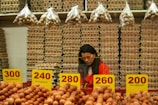 A person stands behind a counter in a market filled with shelves of stacked egg cartons. Baskets of eggs are in the foreground, with brightly colored price labels displayed prominently. Small bags of quail eggs hang from above the stacks of cartons.