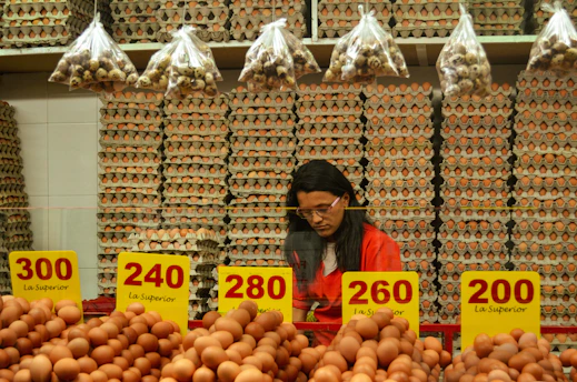 A person stands behind a counter in a market filled with shelves of stacked egg cartons. Baskets of eggs are in the foreground, with brightly colored price labels displayed prominently. Small bags of quail eggs hang from above the stacks of cartons.