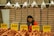 A person stands behind a counter in a market filled with shelves of stacked egg cartons. Baskets of eggs are in the foreground, with brightly colored price labels displayed prominently. Small bags of quail eggs hang from above the stacks of cartons.