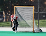 A young athlete wearing a red helmet and black and red uniform is actively guarding a lacrosse goal on a sports field. The player is positioned in front of the net, holding a lacrosse stick. The background shows a chain-link fence and some trees.
