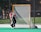 A young athlete wearing a red helmet and black and red uniform is actively guarding a lacrosse goal on a sports field. The player is positioned in front of the net, holding a lacrosse stick. The background shows a chain-link fence and some trees.