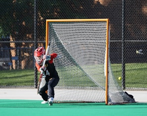 A young athlete wearing a red helmet and black and red uniform is actively guarding a lacrosse goal on a sports field. The player is positioned in front of the net, holding a lacrosse stick. The background shows a chain-link fence and some trees.
