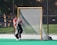 A young athlete wearing a red helmet and black and red uniform is actively guarding a lacrosse goal on a sports field. The player is positioned in front of the net, holding a lacrosse stick. The background shows a chain-link fence and some trees.