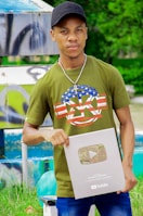 A person wearing a green t-shirt with a decorative design and a black cap is holding a YouTube silver play button. The background features a colorful and artistic mural, as well as some greenery.