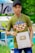 A person wearing a green t-shirt with a decorative design and a black cap is holding a YouTube silver play button. The background features a colorful and artistic mural, as well as some greenery.