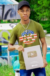 A person wearing a green t-shirt with a decorative design and a black cap is holding a YouTube silver play button. The background features a colorful and artistic mural, as well as some greenery.