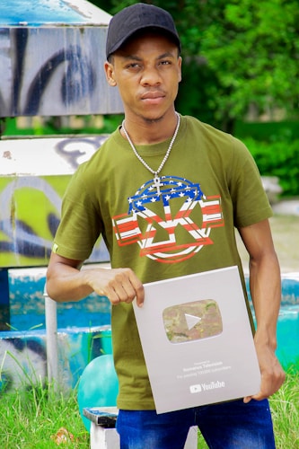 A person wearing a green t-shirt with a decorative design and a black cap is holding a YouTube silver play button. The background features a colorful and artistic mural, as well as some greenery.