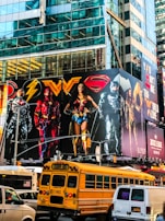 A large, colorful billboard featuring several superheroes in iconic costumes is mounted on the side of a tall, glass-covered building. Below the billboard, traffic includes a yellow school bus and a van. The urban setting is vibrant and lively, with reflections of the city’s architecture visible on the building’s surface.
