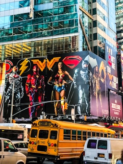 A large, colorful billboard featuring several superheroes in iconic costumes is mounted on the side of a tall, glass-covered building. Below the billboard, traffic includes a yellow school bus and a van. The urban setting is vibrant and lively, with reflections of the city’s architecture visible on the building’s surface.
