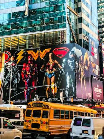 A large, colorful billboard featuring several superheroes in iconic costumes is mounted on the side of a tall, glass-covered building. Below the billboard, traffic includes a yellow school bus and a van. The urban setting is vibrant and lively, with reflections of the city&rsquo;s architecture visible on the building&rsquo;s surface.