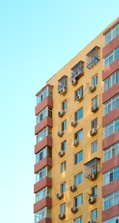A multi-story residential building with a yellow facade, featuring numerous windows. Each window is equipped with an air conditioning unit. The building is accented with red horizontal lines separating each floor. The sky is clear and light blue in the background.