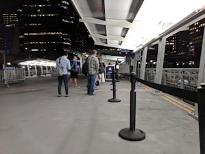 A group of people stand in a line on a platform that appears to be part of a ferry terminal or similar transportation hub. The setting is predominantly urban with tall buildings visible in the background, and it's nighttime with artificial lighting illuminating the area. Metallic railings and barriers are present, and there is a roof overhead.