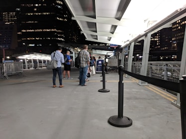 A group of people stand in a line on a platform that appears to be part of a ferry terminal or similar transportation hub. The setting is predominantly urban with tall buildings visible in the background, and it's nighttime with artificial lighting illuminating the area. Metallic railings and barriers are present, and there is a roof overhead.