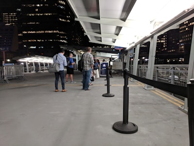 A group of people stand in a line on a platform that appears to be part of a ferry terminal or similar transportation hub. The setting is predominantly urban with tall buildings visible in the background, and it's nighttime with artificial lighting illuminating the area. Metallic railings and barriers are present, and there is a roof overhead.